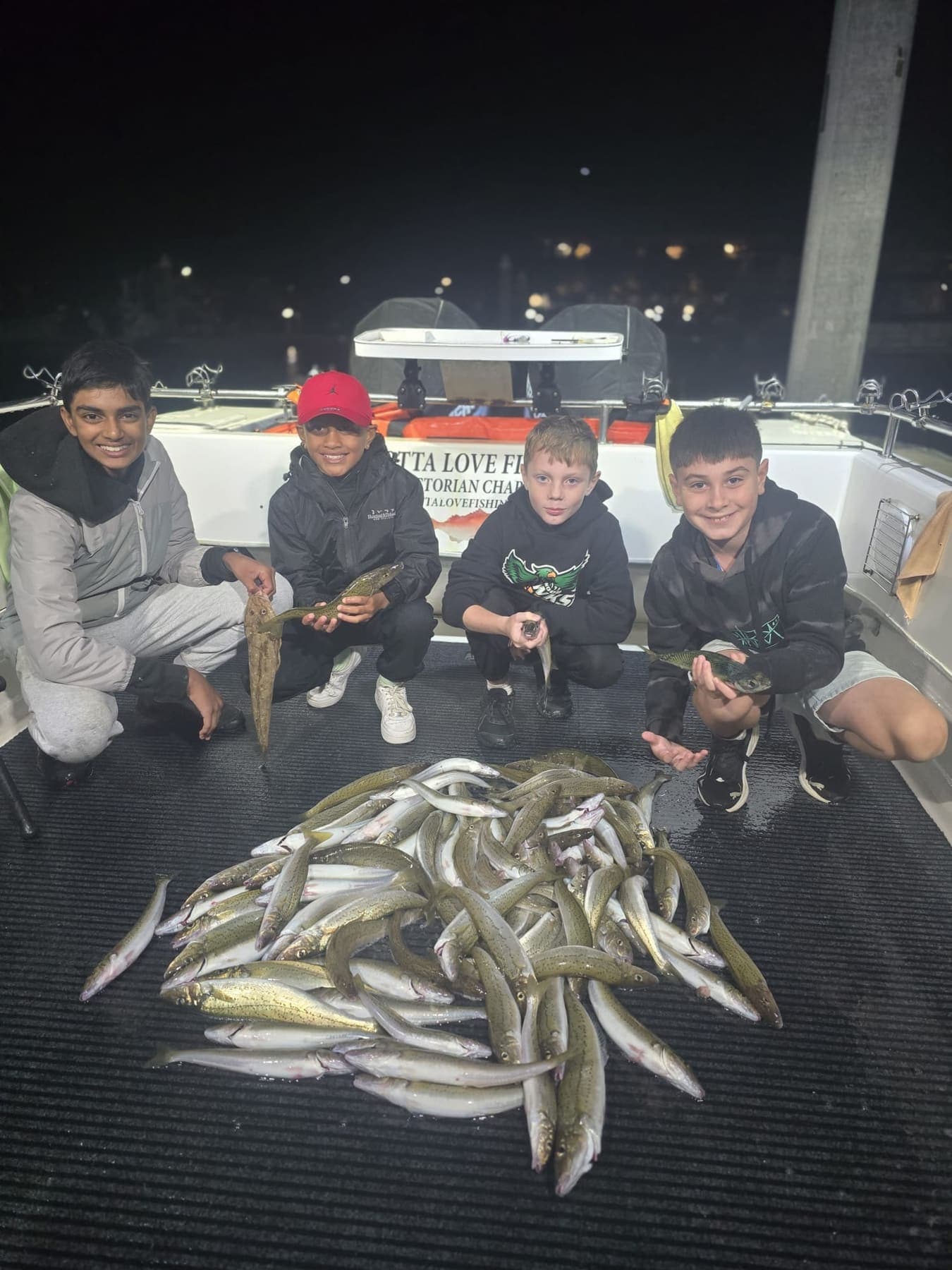 Four kids on the deck at night holding up fish behind a big pile of whiting