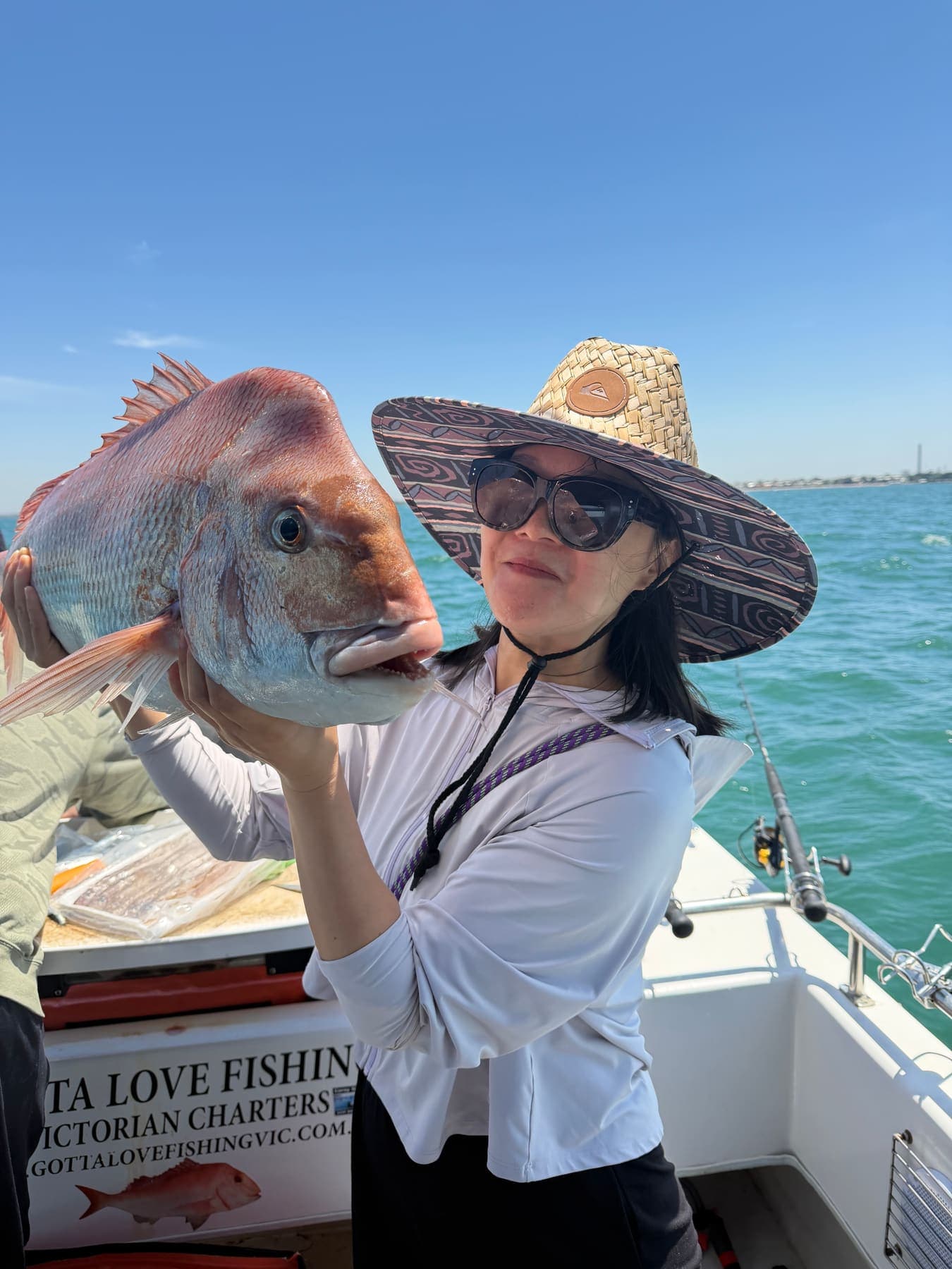 A guest in a wide straw hat holding a big snapper up beside her face on the boat